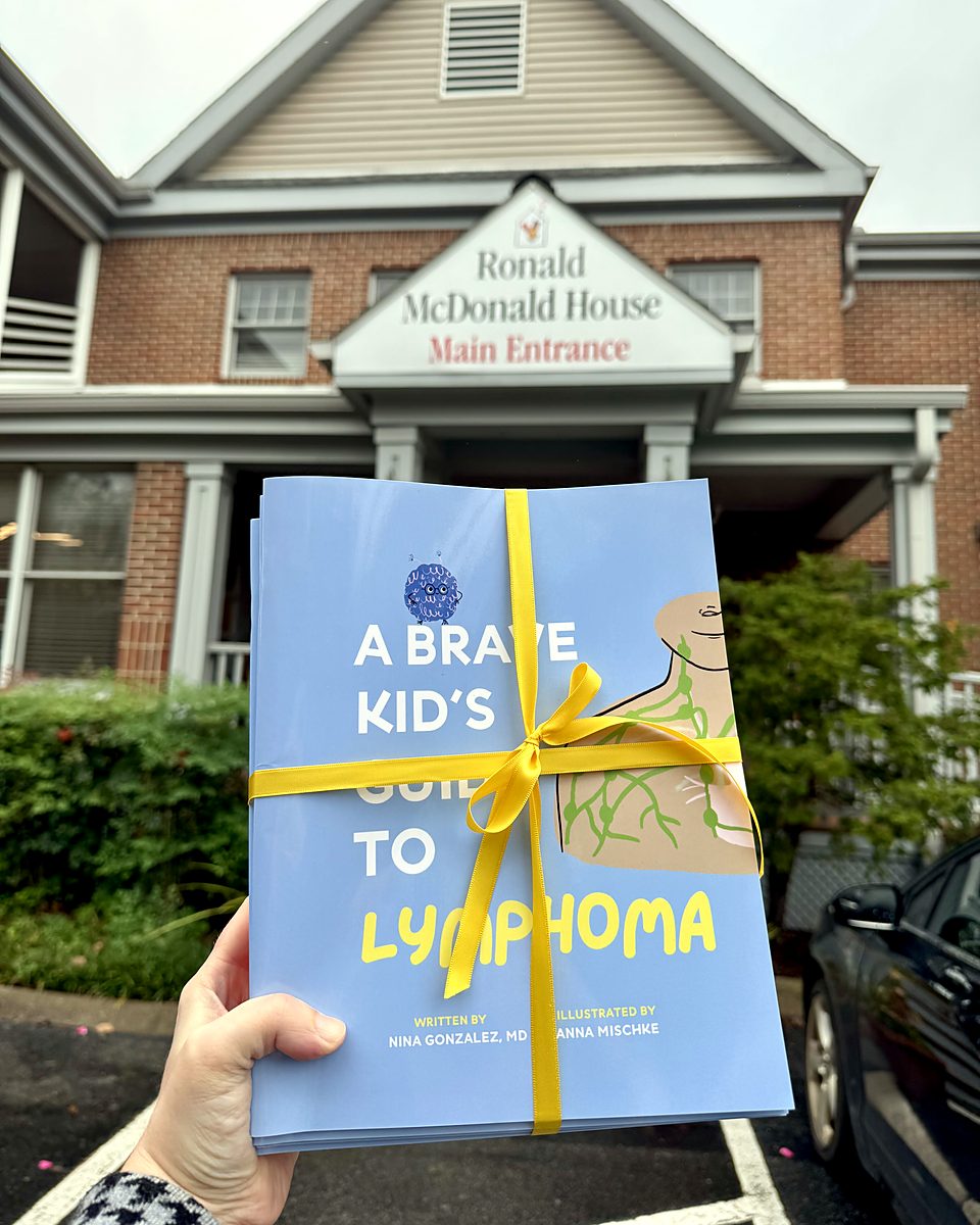 A stack of A Brave Kid's Guide to Lymphoma, tied with a yellow ribbon, held up in front of a Ronald McDonald House entrance