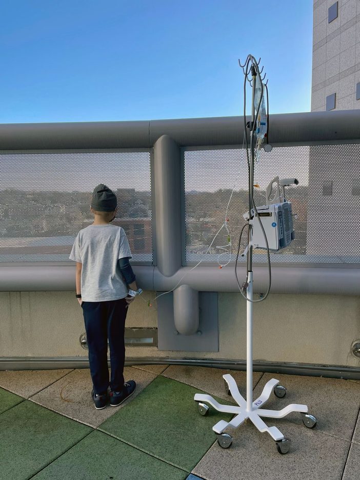 A young boy in a beanie, seen from behind, stands at a hospital rooftop railing next to his IV pole
